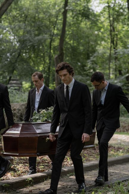 Three men in formal dark suits participate in a funeral procession outdoors, carrying a polished wooden coffin on their shoulders along a gravel path surrounded by tall, leafy green trees. The men are